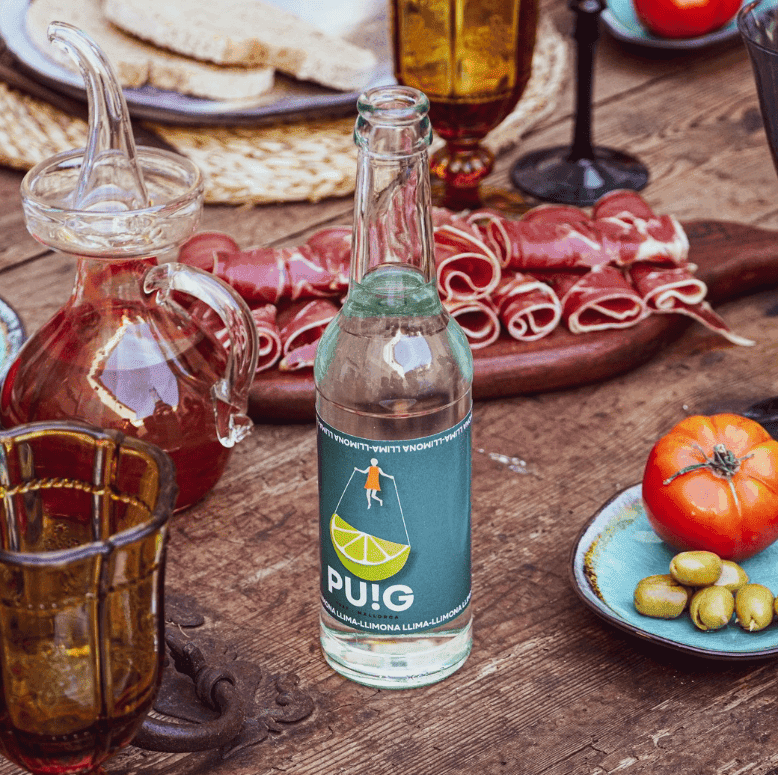 Refresco de lima-limón sobre mesa junto a embutidos, aceite y tomate