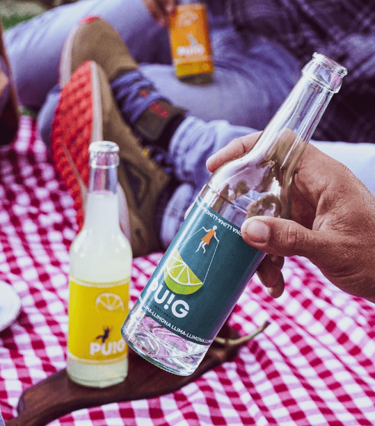 Mano sujetando botella de refresco de lima-limón mientras personas disfrutan de un picnic  