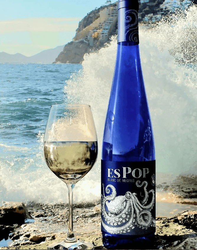 Botella de vi ES POP BLANC DE NEGRES al costat d'una copa de vi blanc, amb una ona de mar de fons. L'ampolla i la copa estan posades sobre una roca al costat del mar, creant una escena fresca i natura