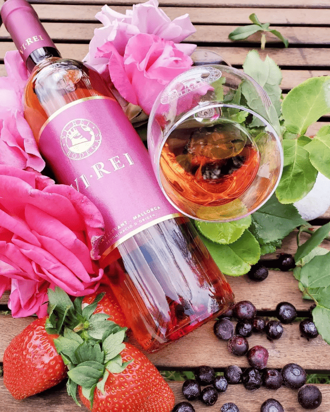 Bottle of Vi Rei Rosé wine lying down, accompanied by a glass of rosé wine with the Vi Rei Winery logo engraved on the glass. The scene is adorned with red and pink fruits around the bottle and glass, creating an elegant atmosphere.