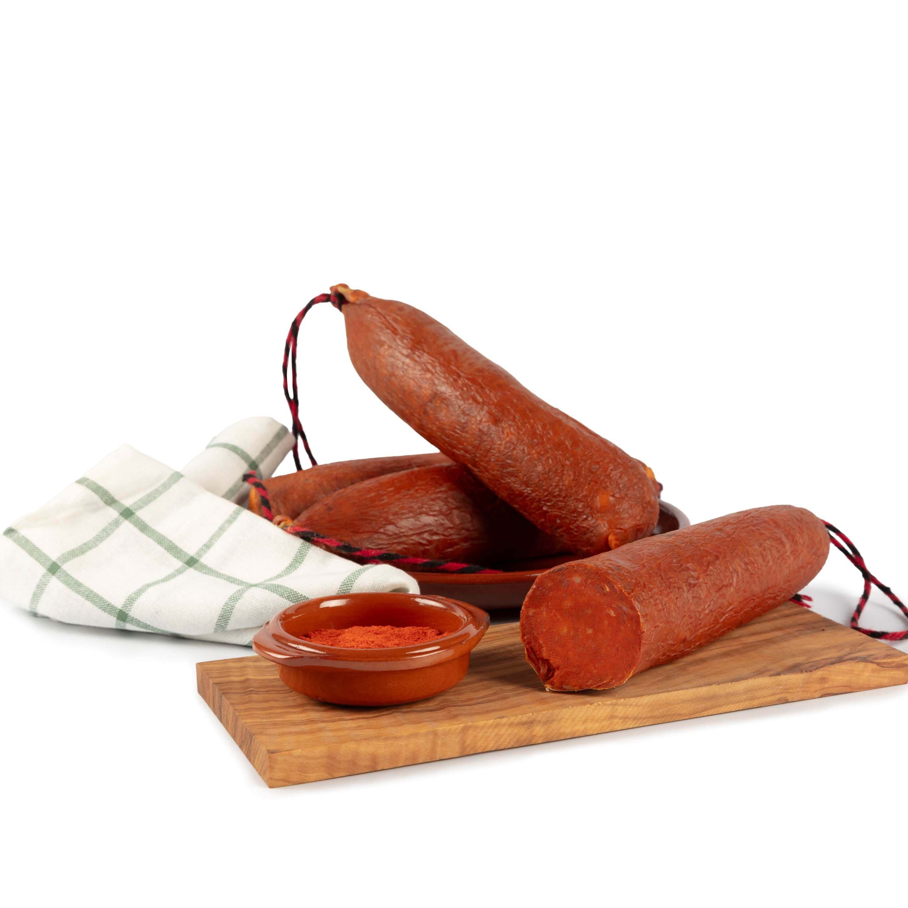 Ganzes Stück und Stück schwarzes Schweinefleisch-Sobrasada aus Mallorca, präsentiert auf einem Brett mit Paprika. Konsumvorschlag und tatsächliches Erscheinungsbild des Produkts.