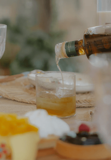 Image of a table with plates of food and, in the center, a glass of ice while pouring Mallorcan herb liqueur "Dos Perellons"
