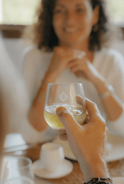 Bild einer Person, die ein Glas mallorquinischen Kräuterlikör in der Hand hält, hinter dem Glas befindet sich eine unscharfe Frau.